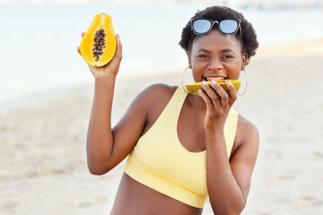Beautiful African girl sitting on the sand, smiling and eating papaya