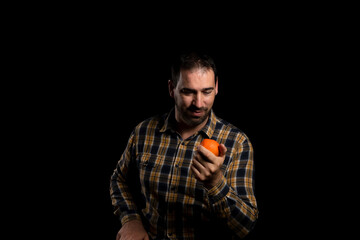 Obraz premium Young bearded man holding and offering an orange on the palm of his hand, isolated on black studio background. Copy space