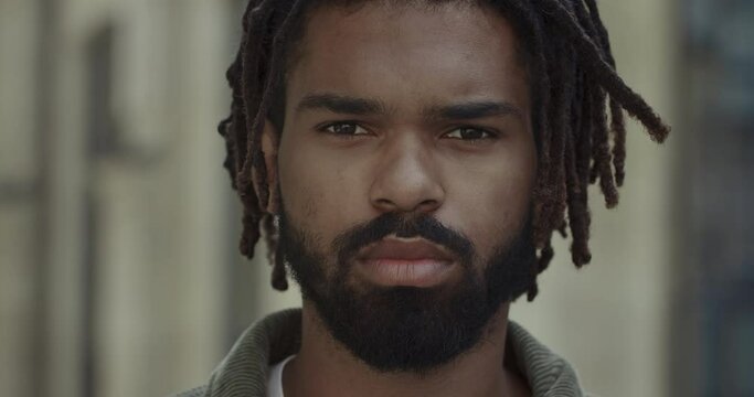 Close up of serious man with beard and moustache looking to camera. Portrait of young guy with dreadlocks standing at city street. Concept of lifestyle