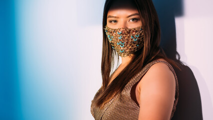 Oriental female beauty. New normal fashion. Party look. Woman in glossy evening dress and golden chain turquoise beads face mask posing on neutral gradient background.