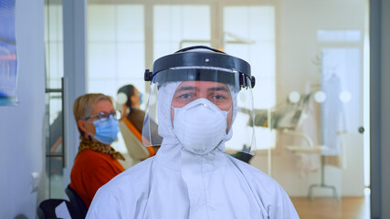 Obraz na płótnie Canvas Close up of tired doctor in stomatological office looking on camera wearing overall and face shield sitting on chair in waiting room clinic. Concept of new normal dentist visit in coronavirus outbreak