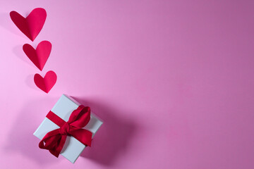 WHITE GIFT WITH RED RIBBON AND PAPER RED HEARTS ON PINK BACKGROUND