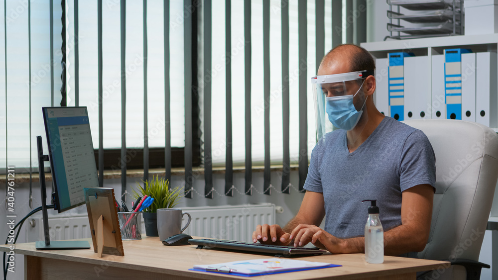Poster man with protection mask and visor applying sanitizer gel rubbing hands before working at computer. 