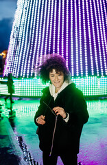Attractive woman in warm clothes stands at the evening square on a background of big christmas tree.