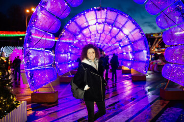 Curly hair smiling young woman in a coat outdoors with Christmas lights and decoration in the background.