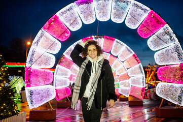 Curly hair smiling young woman in a coat outdoors with Christmas lights and decoration in the background.