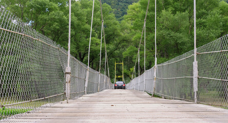 A black car is crossing hanging bridge with wooden planks over the river