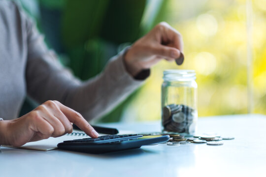 Closeup image of a woman putting coins in a glass jar and calculating on calculator for saving money and financial concept