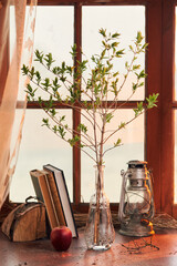 spring twig in a transparent glass vase with a kerosene lamp and books on the background of the window
