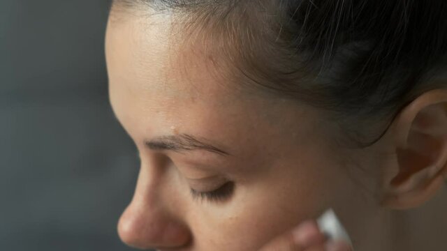 Woman is wiping her eyebrows from paint with a cotton pad dipped in water looking at mirror. Beauty procedure at home, face closeup view. Modern trendy styling, correction, coloring of eyebrows.