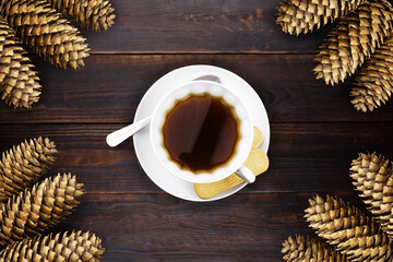 White cup with coffee, spoon and saucer, cookies on brown wooden plank surface with fir cones in...