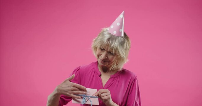 Close up portrait of cheerful happy senior Caucasian woman wearing birthday hat isolated posing on pink background in studio smiling and holding in hands cute envelope holiday card. positive emotions.