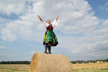 Naklejka na ściany i meble A pretty happy young girl dressed in Polish traditional national folk dress standing in the field on a harvest straw bales under a blue sky
