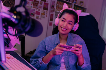 Young playful Asian woman sitting in front of her computer workstation and smiling at the screen with a microphone in foreground. Beautiful asian woman working from her home studio and playing game. © twinsterphoto