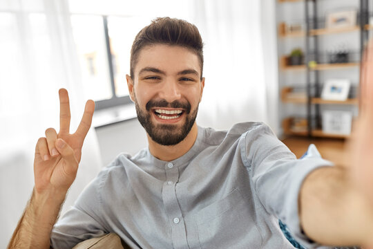 technology, people and lifestyle concept - happy man taking selfie or having video call and showing peace gesture at home