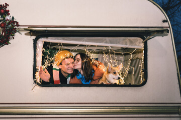 Happy caucasian couple with corgi dog in a caravan or campervan. Woman kissing a man, casual winter...