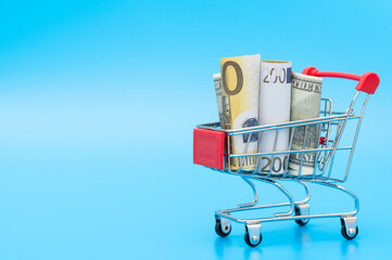 Shopping cart with American and European banknotes on blue background. Copy space for text