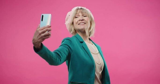 Happy cheerful Caucasian senior cute pretty woman wearing beret and green jacket standing isolated on pink background using cellphone taking selfie photos on smartphone smiling while posing. portrait