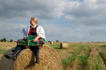 Naklejka na ściany i meble A pretty happy young girl dressed in Polish traditional national folk dress sitting in the field on a harvest straw bales under a blue sky