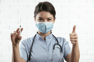 Beautiful female doctor with medical mask. Portrait of nurse with syringe showing thumbs up.