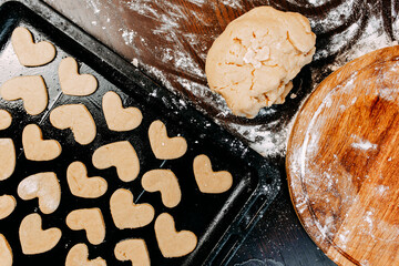 Cookies in shape of heart for the Saint Valentine's Day. Dough, flour, baking pan, round wooden cutting board and rolling pin on the table.
