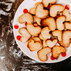 Cookies in shape of heart for the Saint Valentine's Day. Dough, flour, baking pan, round wooden cutting board and rolling pin on the table. Cookies on the plate.