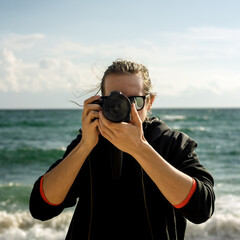 Male photographer wearing black jacket holding a DSLR camera with sea and cloudy sky in the background