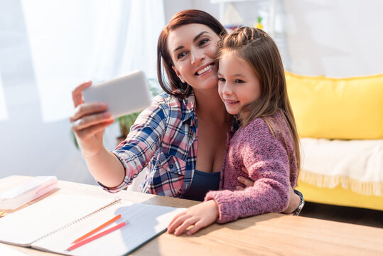 Cheerful mother and daughter taking selfie at desk with copy book on blurred foreground
