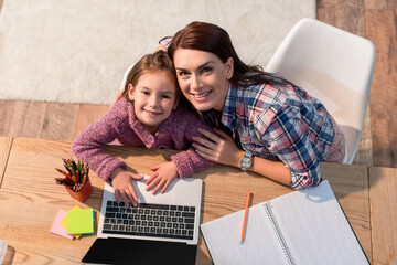 Top view of smiling mother and daughter looking at camera while sitting at desk with laptop and stationery