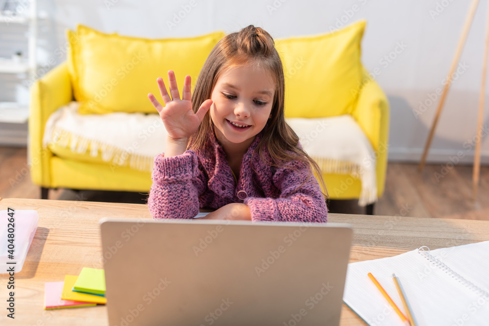 Wall mural smiling girl with waving hand looking at laptop on desk with stationery on blurred foreground