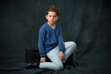 Obraz premium teen boy in a blue sweater and blue trousers posing while sitting in the studio on a gray fabric background