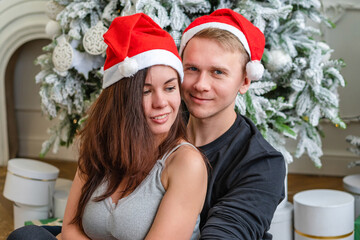 A loving couple of man and woman embrace next to a Christmas tree, dressed in Santa hats.