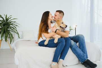 Cheerful married couple is playing with dog at home. They are sitting on sofa and stroking the animal. The man and woman are smiling