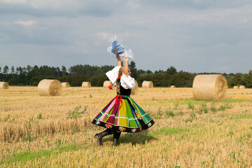 Blonde pretty young smiling girl dressed in Polish national folk dress dancing and spinning around in the field of harvest straw bales