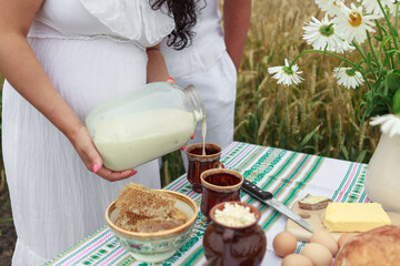  Hands of a pregnant girl in a white dress pour fresh milk from a can into a mug. honey, eggs, butter on the table. Wheat field. High quality photo