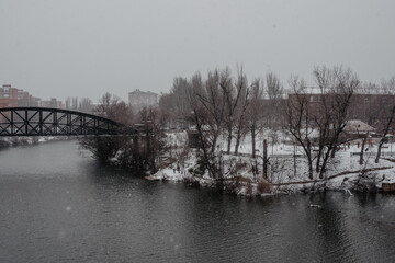 Paisaje de un río con orillas cubiertas de nieve en un día de invierno. Caída de nieve. Puente de metal negro