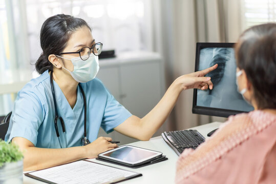 Asian female doctor in medical mask examining and pointing to radiological chest X-ray film on monitor computer with Elderly woman patient