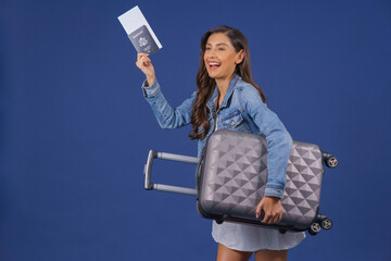 Happy young Asian woman white dress with jean jacket  holding passport with flying tickets while standing with a suitcase on blue color background, Travel concept