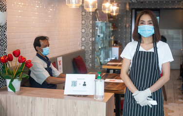 Asian waitress woman wearing face masks and holding an infrared forehead thermometer to check body temperature for virus symptoms of customers before entering the restaurant ( coffee shop ). - Powered by Adobe
