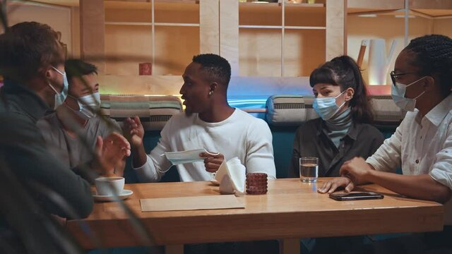 Coronavirus time. Group of multiracial people making their friend wear a protective mask while sitting at cafe. Company of people explaining to their friend why he need to wear a mask at the pandemic
