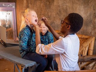 African American woman wearing white coat dentist examines blonde boy in village clinic. child's brothers are waiting for their turn and are worried about baby