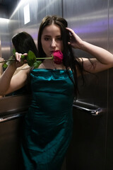 sexy confident brunette woman in elegant aquamarine colored dress with red rose in grey elevator
