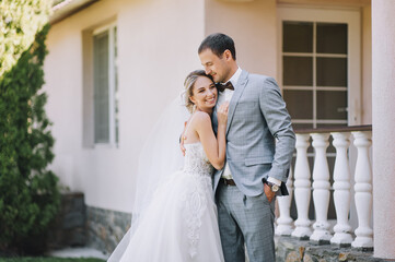 Naklejka premium Stylish groom in a gray checkered suit and a cute blonde bride in a white lace dress with a long veil are hugging in the park near the old house. Wedding portrait of smiling newlyweds.