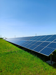 A row of solar panels stand on a green field, against the blue sky. A symbol of clean energy, concern for the environment, a concept.