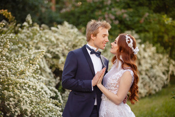 Young Bride and Groom couple in a blooming garden. Tender holding each other. Spring wedding. Redhead girl with long hairs. Young family outdoor image near blooming bush of spirea. Love and tenderness