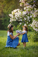 Mother and little daughter in blooming lilac garden with lilac bouquet. Mom loves her child. Spring story. Family look romantic similar blue dresses and straw hat. Happy family in beautiful spring day - 404861847