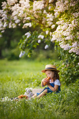 Little girl weared in romantic rustic look on picnic in a blooming lilac garden. Spring story. Romantic rustic look with blue dress and straw hat. Happy girl in beautiful spring day. 