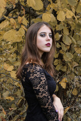 Girl with dark makeup and in a black lace dress posing on a background of leaves. Gothic shooting. Witchcraft and vampire style. Portrait.