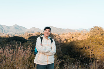 Naklejka na ściany i meble Smiling Asian young man in Long-sleeved shirt and grey hat hiking Stand with arms folded at mountain peak above clouds Hiker outdoor. Lamphun Province, Northern Thailand Province in the morning.