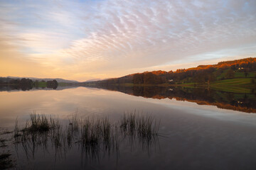 Peaceful dusk falls across Esthwaite Water with reflections in the water of buildings, trees and colourful clouds in a mackerel sky, Lake District, UK